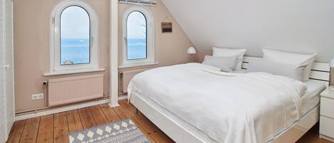 A bedroom with a large bed, two windows with a view of the sea, a radiator, and a wooden floor.