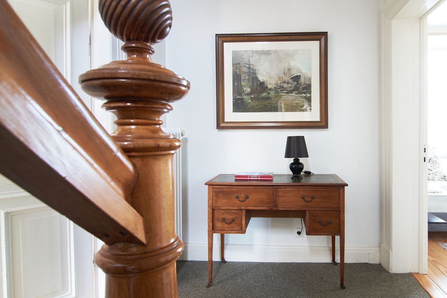 A wooden staircase with a balustrade, a desk with drawers, a framed painting, and a lamp.