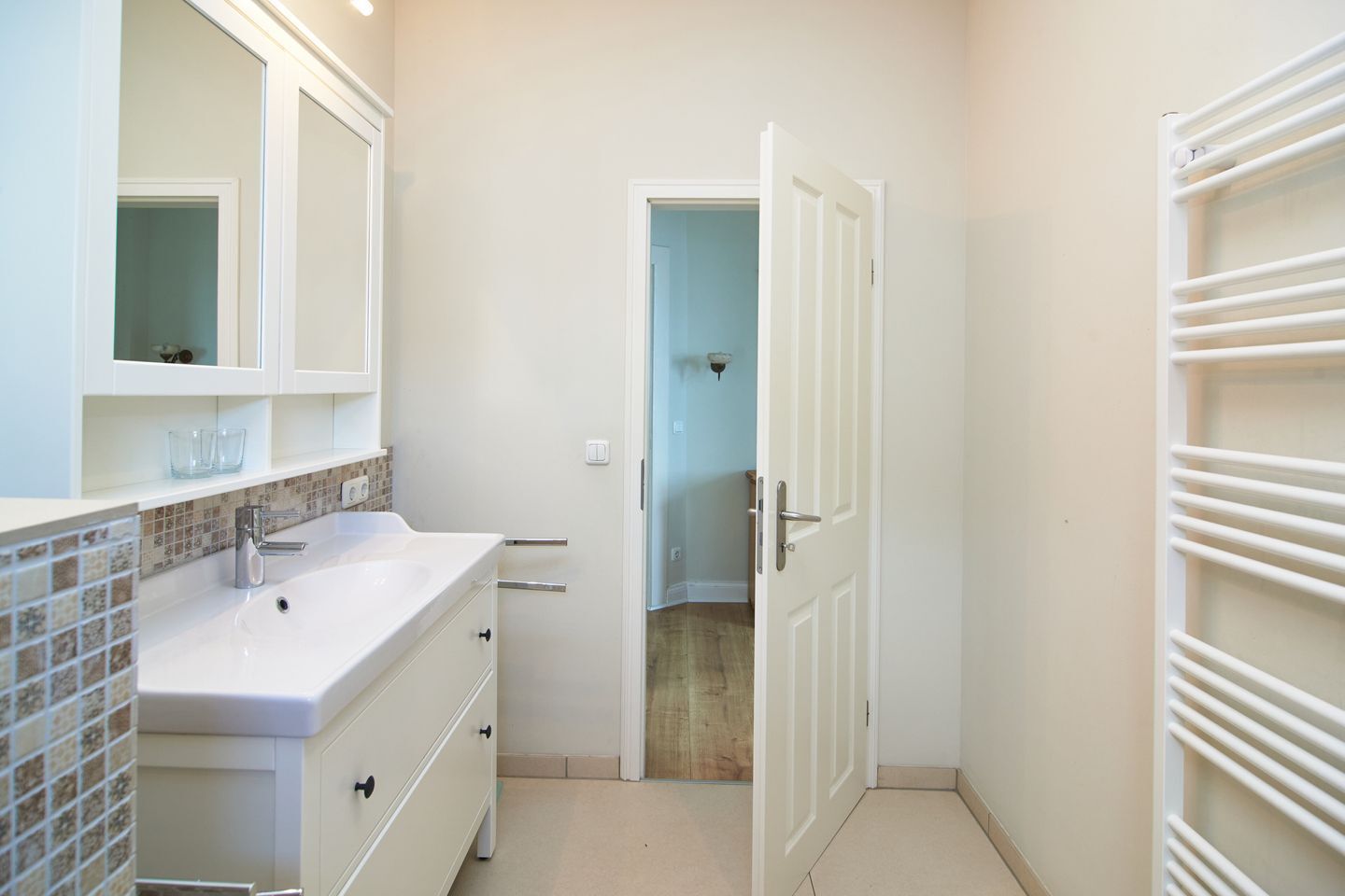 Un baño con un lavabo blanco, espejo y salpicadero de azulejos. Hay una puerta que da a otra habitación.
