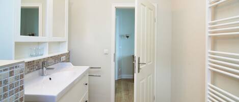 Un baño con un lavabo blanco, espejo y salpicadero de azulejos. Hay una puerta que da a otra habitación.