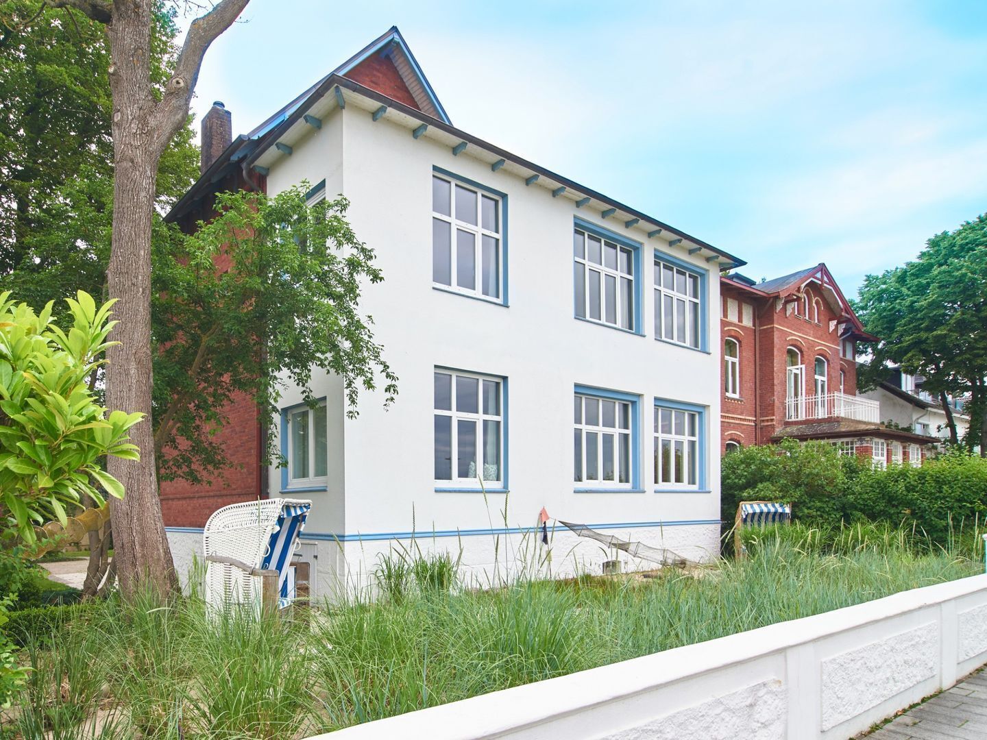 A two-story building with a white facade and blue trim, surrounded by greenery and a white fence.