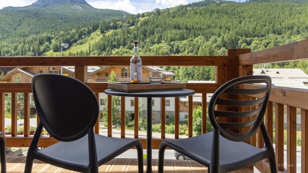 Ein Balkon mit zwei schwarzen Stühlen, einem kleinen runden Tisch und einer Flasche Alkohol. Der Balkon bietet Blick auf ein bewaldetes Gebirge.