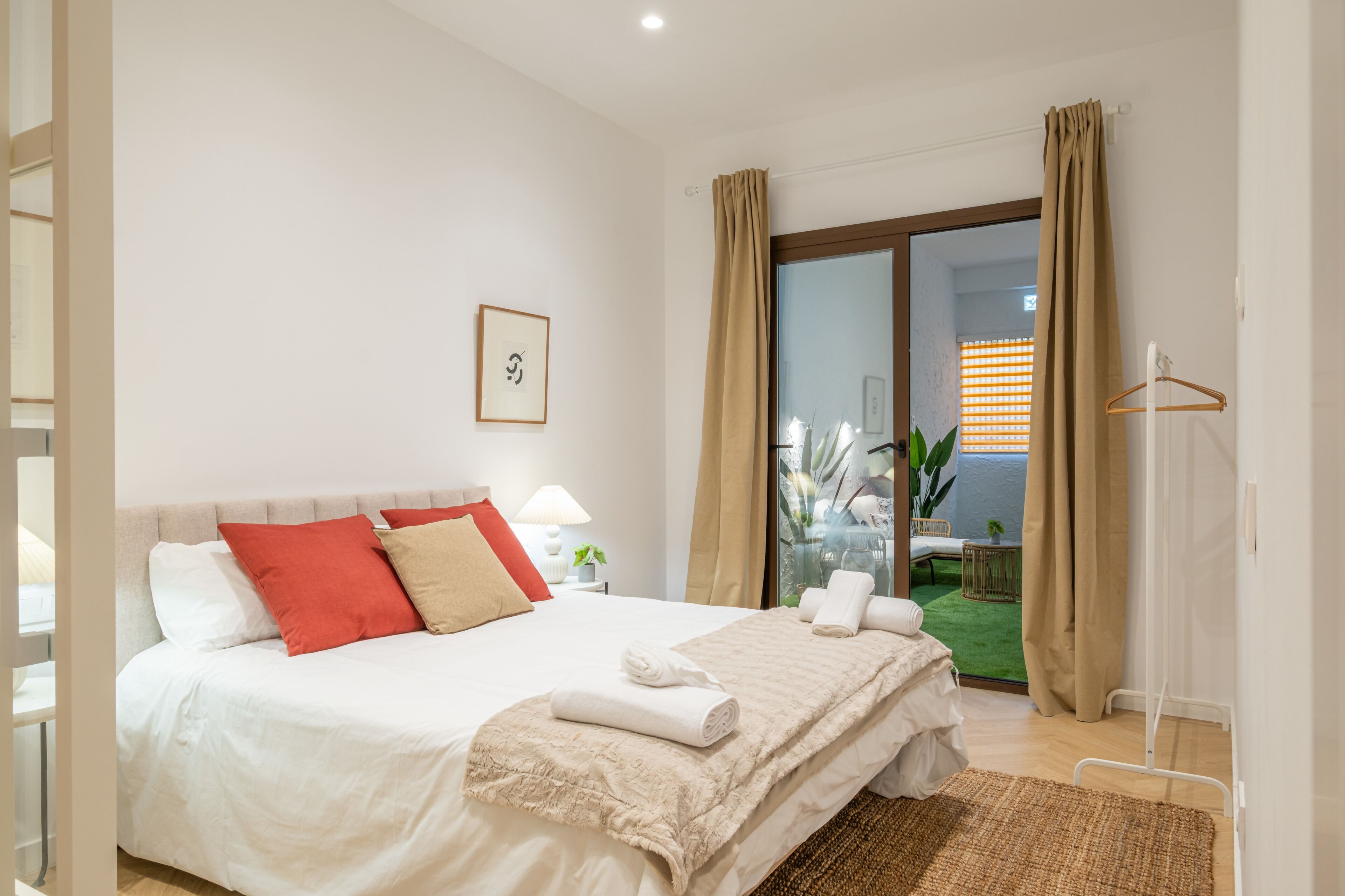 Habitación de hotel con cama, mesita de noche con lámpara, ventana con cortinas y vistas al exterior.