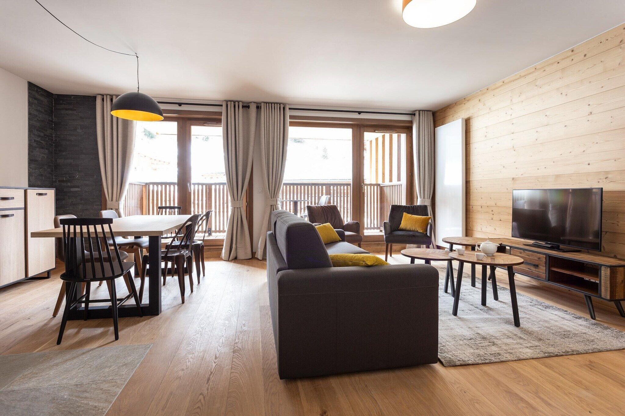 Uma sala de estar moderna com sofá, mesa de jantar, cadeiras, televisão e varanda com cortinas.