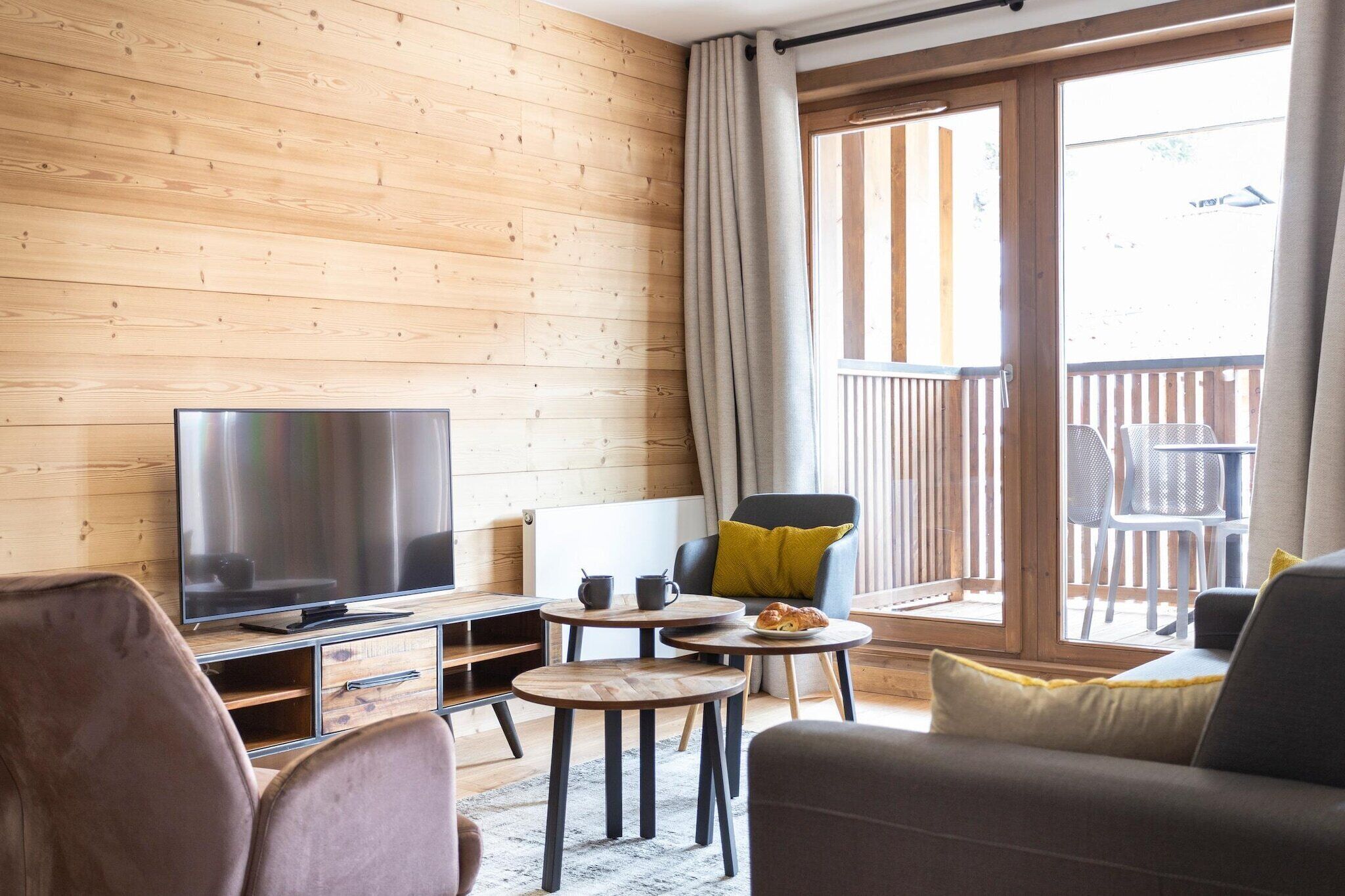 A cozy room with a wooden wall, a flat-screen TV, a sofa, a dining table with chairs, and a balcony with a railing.