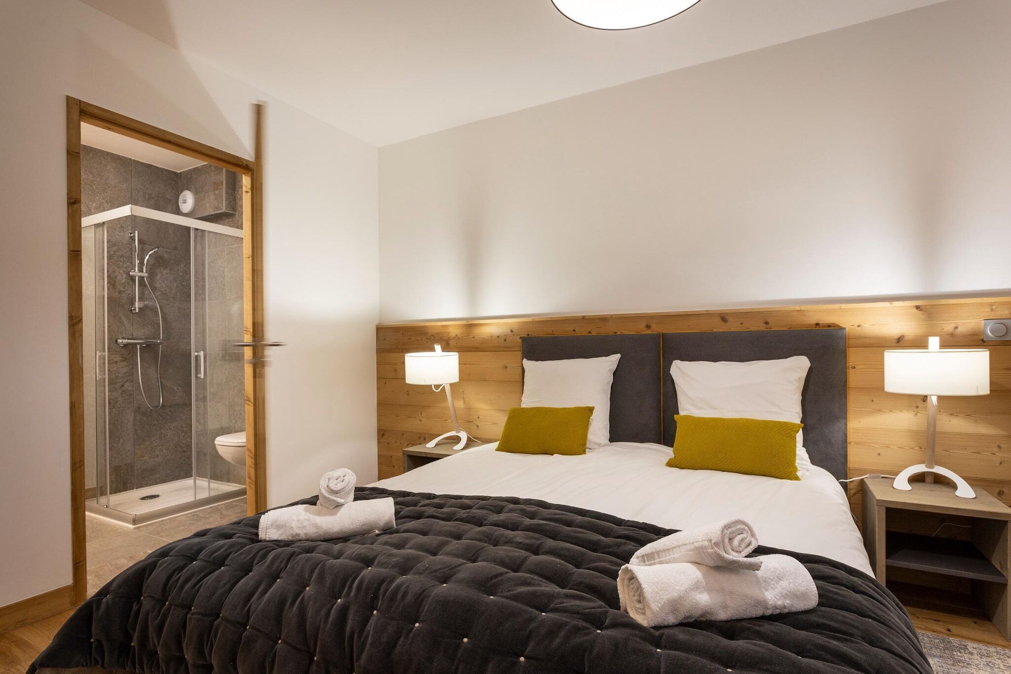 A modern hotel room with a large bed, two bedside lamps, a wooden headboard, and a glass-enclosed shower area.