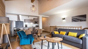 Ein modernes Wohnzimmer mit grauem Sofa, blauem Sessel und Holztisch. Im Hintergrund ist eine Küche mit Essbereich erkennbar.