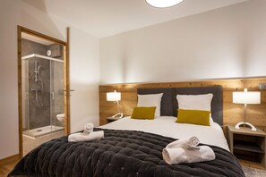 Une chambre d’hôtel moderne avec un grand lit, deux lampes de chevet, une tête de lit en bois et une cabine de douche en verre.