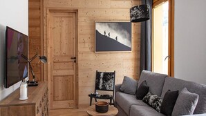 A cozy living room with a wooden door, a grey sofa, a black and white patterned chair, a round wooden table, and a mounted picture of hikers on a mountain. - Apartment in Serre Chevalier Ski Resort (VAUJANY)
