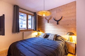 Une chambre à coucher avec un grand lit, deux lampes de chevet, une fenêtre avec des rideaux et un mur en bois orné d’une tête d’animal fixée au mur.