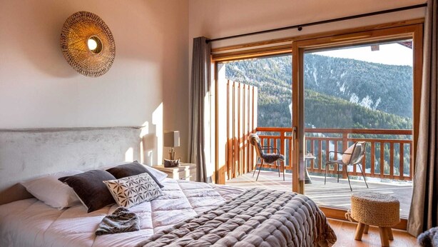 A bedroom with a large bed, a balcony with a view of a forested mountain, and a woven wall light.