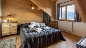 A bedroom with a wooden ceiling, a bed with a dark bedspread, a bedside lamp, a radiator, and a window with curtains.