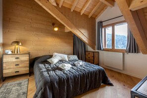 A bedroom with a wooden ceiling, a bed with a dark bedspread, a bedside lamp, a radiator, and a window with curtains. - Apartment in Alpe d'Huez near Grandes Rousses (VAUJANY)