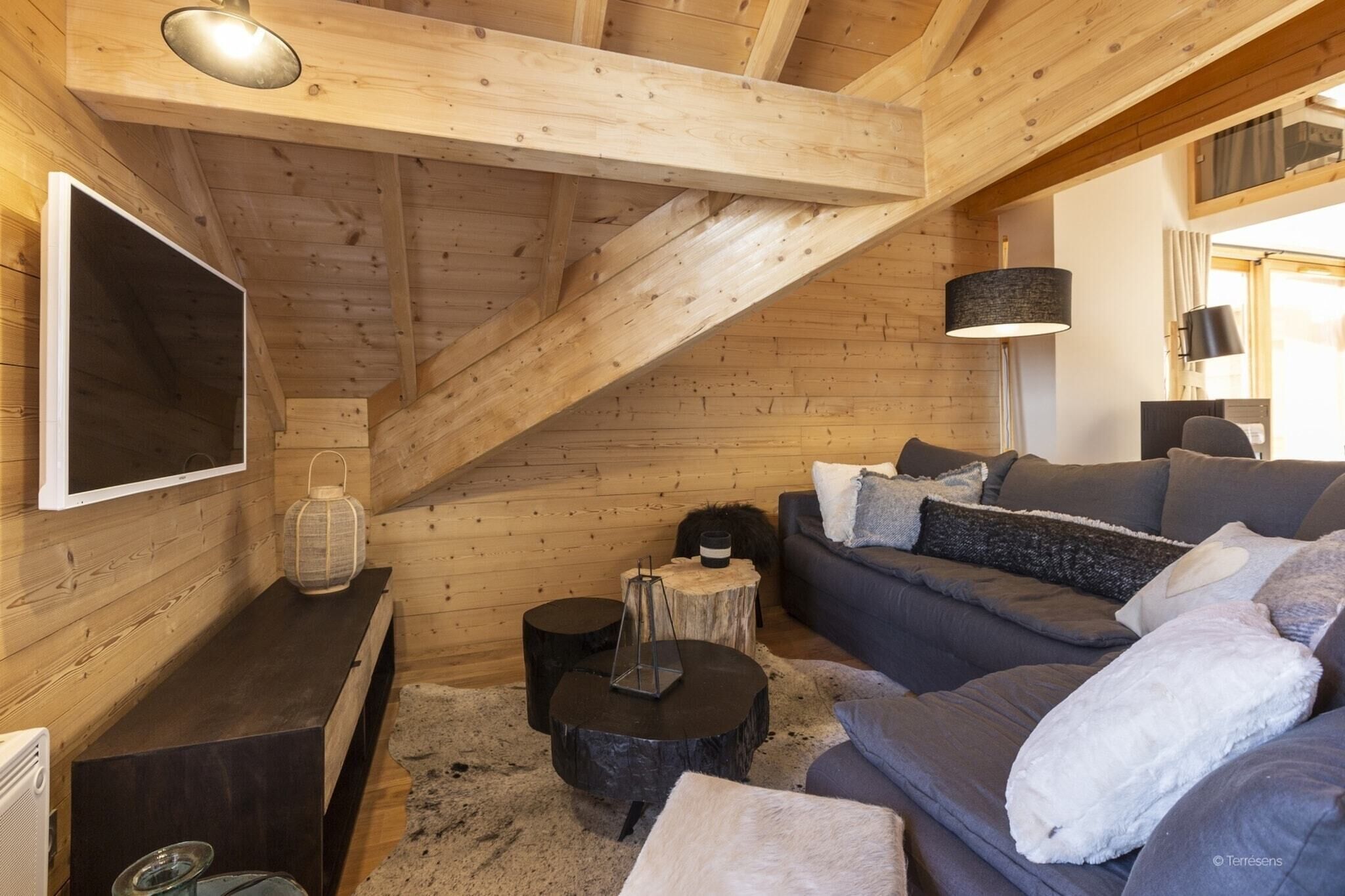 Interior acolhedor de uma cabana, com teto de madeira, um sofá, uma televisão e uma pequena mesa com um candeeiro.