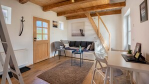 A room with a wooden ceiling, a sofa, a table, a chair, a radiator, and a door. - Guesthouse 'Charming Lütt Hus' with Private Terrace, Shared Garden and Wi-Fi (Damlos)