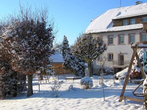 Un espace extérieur enneigé, avec des arbres, une balançoire et un bâtiment doté de nombreuses fenêtres.