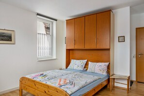 A bedroom with a wooden bed, a window with curtains, a wooden wardrobe, and a small bedside table.