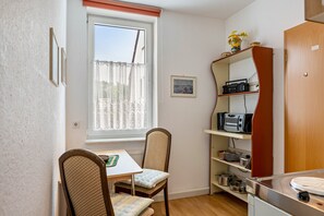 A compact kitchen with a dining table, chairs, a window with curtains, and a shelf with kitchen appliances.