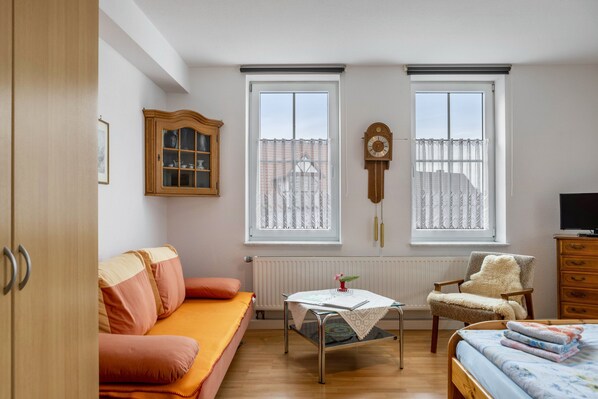 A bedroom with a bed, a sofa, a chair, a table, two windows, a radiator, and a clock on the wall.