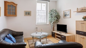 Ein Wohnzimmer mit einem Ledersofa, einem Glastisch, einem Fernseher, einem Holzschrank und einem großen Fenster mit Jalousien.