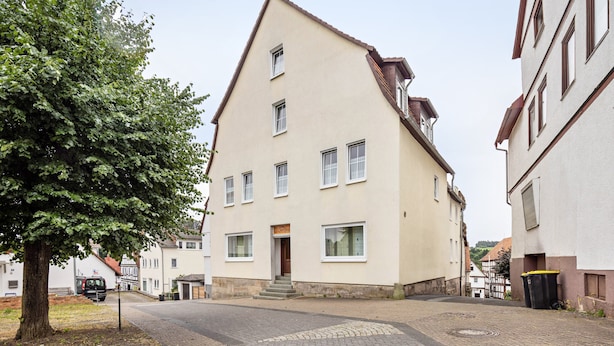 Ein zweistöckiges Gebäude mit einem geneigten Dach, einem zentralen Eingang mit Stufen und mehreren Fenstern.