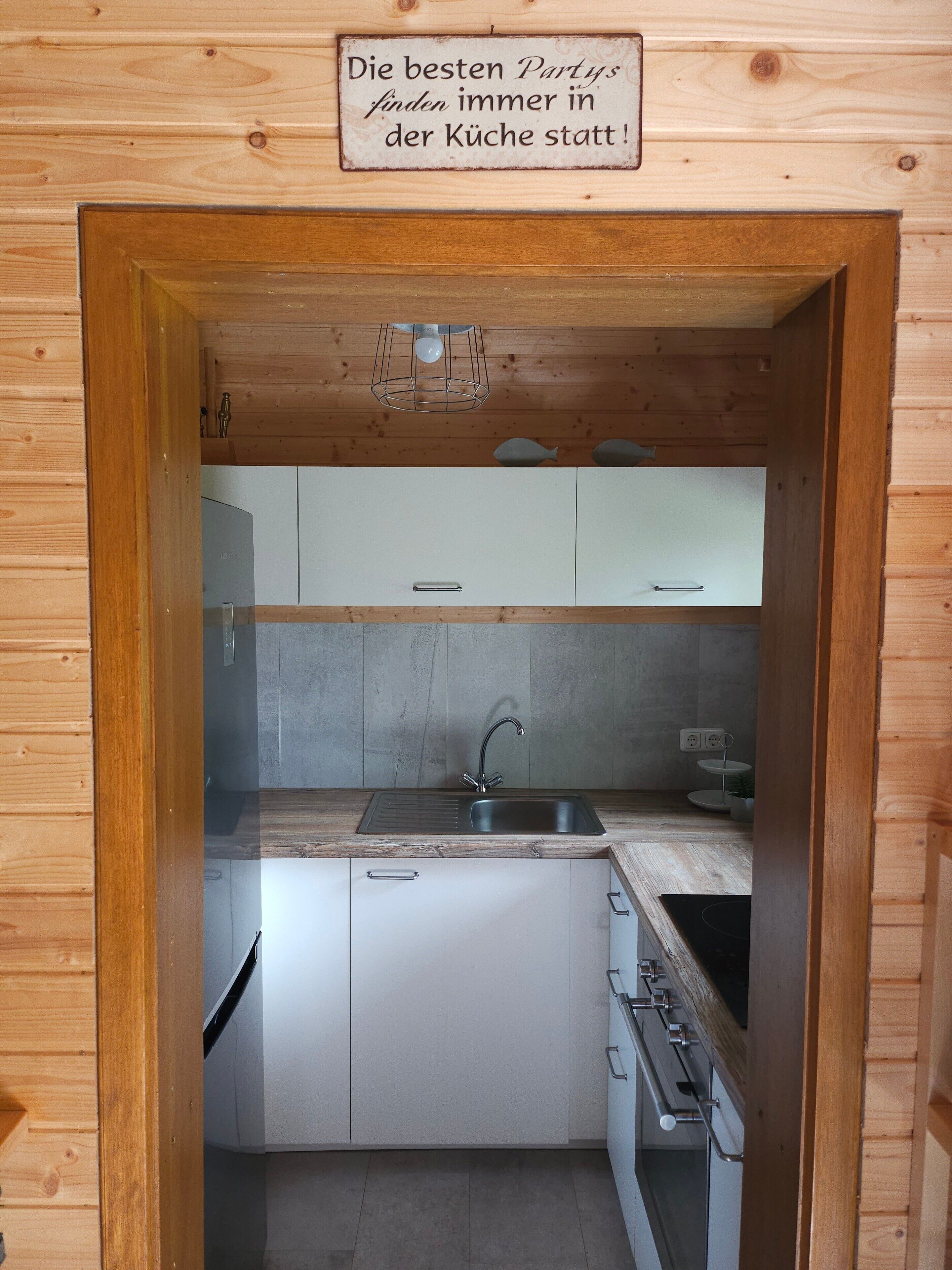 A compact kitchen with wooden walls, a stainless steel sink, white cabinets, and a sign above the door.