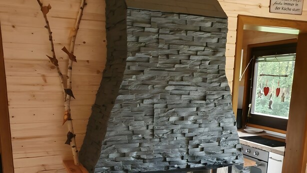 Een open haard van natuursteen met een houten bovenkant, een raam met decoraties en een keuken in de achtergrond.