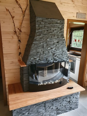 A stone-built fireplace with a wooden mantel, a window with decorations, and a kitchen area in the background. - Holiday Home 'Weiherhüttla' with Lake View, Private Terrace and Garden (Untersteinach)