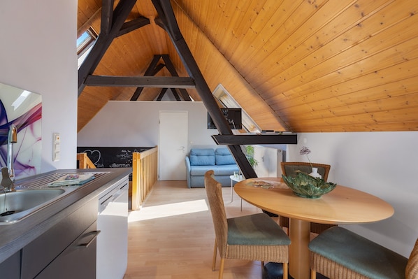 Une cuisine moderne dotée d’un lave-vaisselle intégré, un coin repas avec une table ronde et des chaises en osier, et une chambre mansardée avec un plafond en pente.