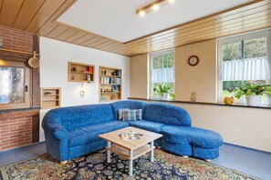Ein Wohnzimmer mit einem blauen Ecksofa, einem Holztisch und einem gemusterten Teppich. Es gibt Regale mit Büchern und dekorativen Gegenständen, einen Kamin aus Ziegelsteinen und ein Fenster mit Blick auf Grünflächen.