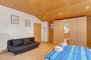 Ein Zimmer mit Holzboden, einer Couch, einem Bett mit blauer Bettdecke, Holzschränken und gerahmten Bildern an der Wand.