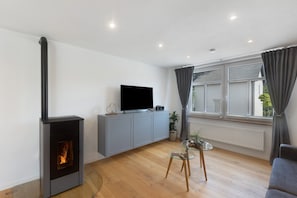 Un salón moderno con chimenea, un televisor de pantalla plana, un armario gris, una mesa de vidrio y un ventanal con cortinas.