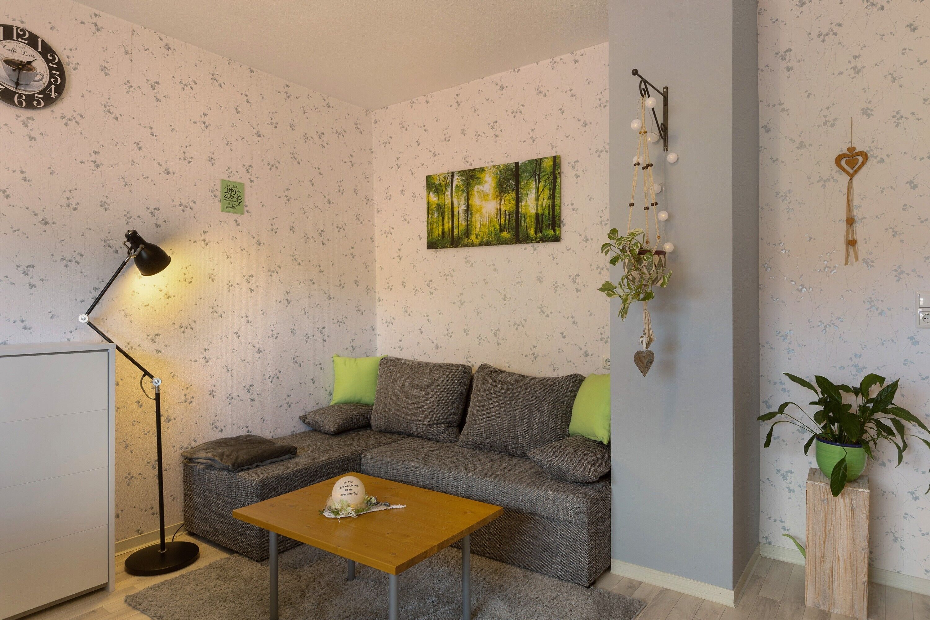 Een woonkamer met een bank, een tafel, een vloerlamp, een klok, een schilderij, wanddecoraties en een plant.