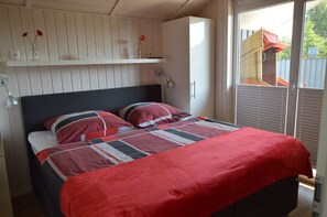A bedroom with a bed, a bedside lamp, a shelf with flowers, and a window with blinds.