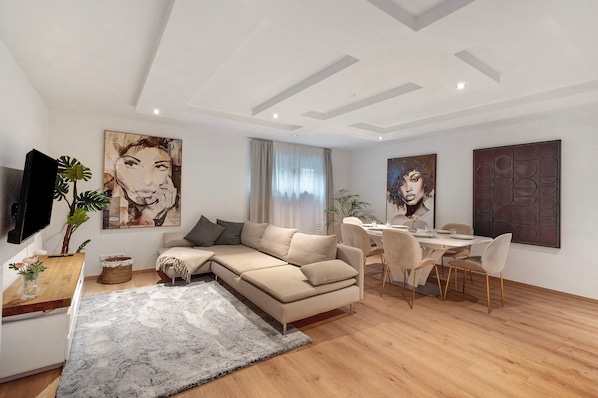 A modern living room with a sofa, dining table, and chairs, a flat-screen TV, and artwork on the walls.