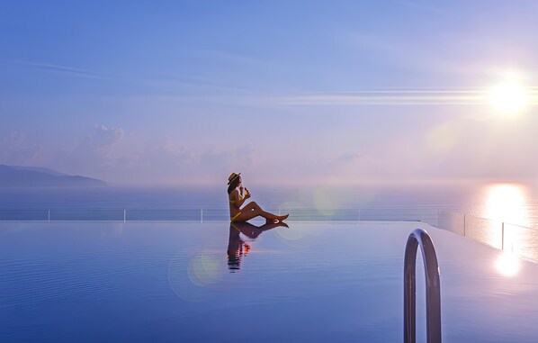 Eine Person sitzt am Poolrand mit freiem Blick auf das Meer und einer hellen Sonne im Hintergrund.