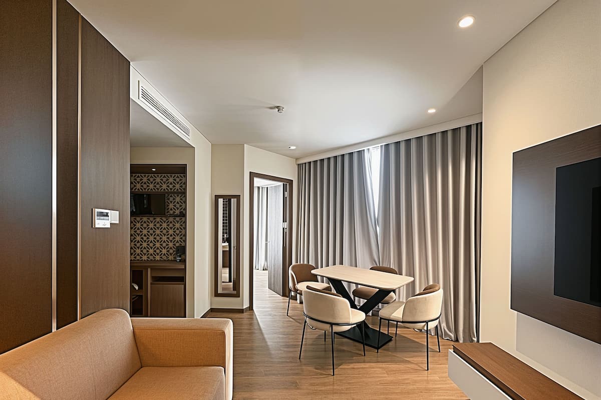 Ein modernes Hotelzimmer mit einer Couch, einem Esstisch mit Stühlen, einem an der Wand befestigten Fernseher und einem großen Fenster mit Vorhängen.