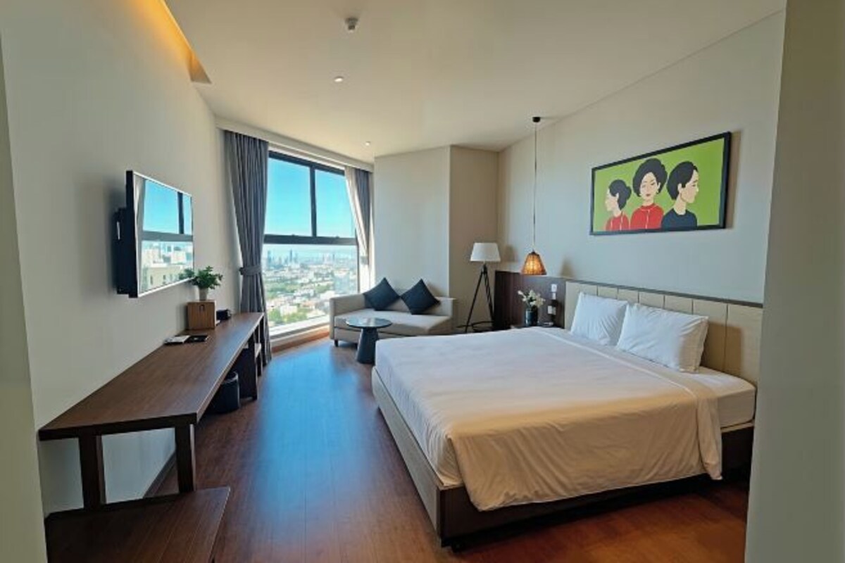 A hotel room with a large bed, a desk, a sofa, a TV, and a window with a city view.