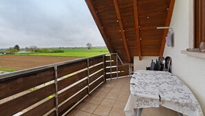 A balcony with a wooden railing, a table with a floral tablecloth, and a view of a green field. - Apartment 'Sigrid' with Shared Garden, Balcony and Wi-Fi (Forchheim)