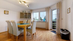 A dining area with a wooden table and chairs, a cabinet with glass doors, a window with curtains, and a trash bin. - Apartment 'Sigrid' with Shared Garden, Balcony and Wi-Fi (Forchheim)