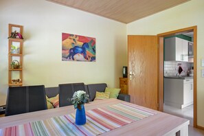 Una sala de estar con un sofá, una mesa baja con un jarrón, un cuadro en la pared y una cocina a la vista a través de un umbral abierto.