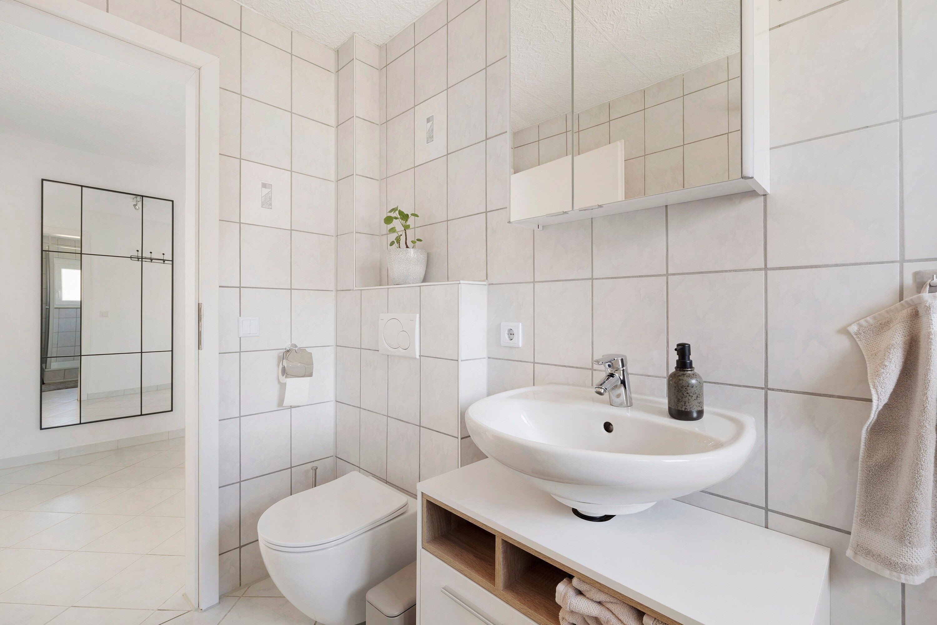 A modern bathroom with white tiles, a white sink, a toilet, a mirror, and a plant.