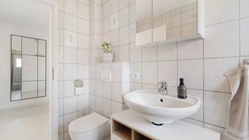 A modern bathroom with white tiles, a white sink, a toilet, a mirror, and a plant.