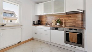 Une cuisine moderne avec des meubles blancs, un pare-feu en bois, un four encastré et une fenêtre donnant sur une maison.