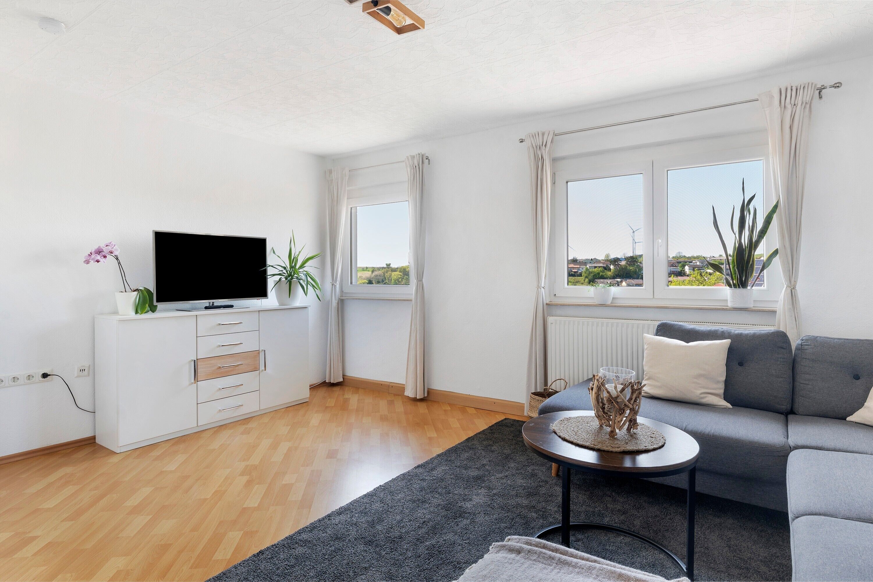 A modern living room with a flat-screen TV, a grey sofa, and a large window with curtains.