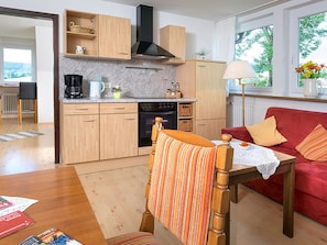 A compact kitchen with wooden cabinets, a stove, and a coffee maker. There is a red sofa with cushions and a wooden chair with a striped cushion. A table with a lace tablecloth is in the foreground. - Apartment 'Meierhof, 38qm' with Shared Terrace, Shared Garden and Wi-Fi (St. Märgen)