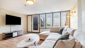A modern living room with a large flat-screen TV, a sofa set, a small table, and a view through large windows.