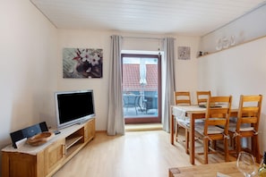 A living room with a TV, wooden furniture, a dining table with chairs, and a balcony with a table and chairs.
