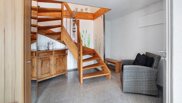 Una escalera de madera que conduce a una zona superior, un sofá de mimbre y un armario de madera con utensilios de cocina.