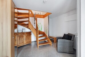 Una escalera de madera que conduce a una zona superior, un sofá de mimbre y un armario de madera con utensilios de cocina.